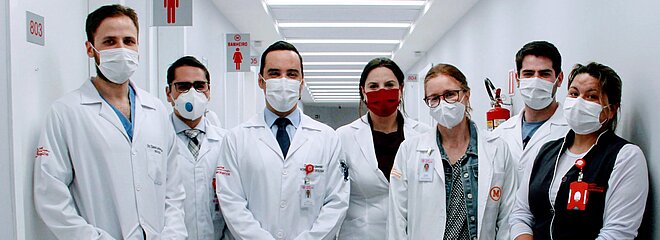 Na imagem, sete pessoas entre homens e mulheres, posam para foto num corredor claro de hospital. As pessoas vestem jalecos e uniformes com símbolo do Mackenzie e estão usando máscaras