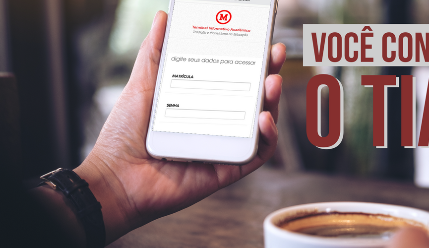 A foto mostra o braço e a mão de um homem usando um relógio segura um celular que aparece a página do aplicativo do TIA. Na mesa na qual ele está se apoiando tem um xícara de café