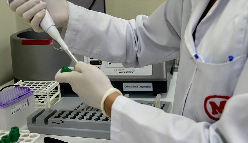 profissional de laboratório com jaleco branco com símbolo do Mackenzie em vermelho, manipulando uma pipeta de laboratório