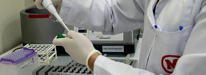 profissional de laboratório com jaleco branco com símbolo do Mackenzie em vermelho, manipulando uma pipeta de laboratório