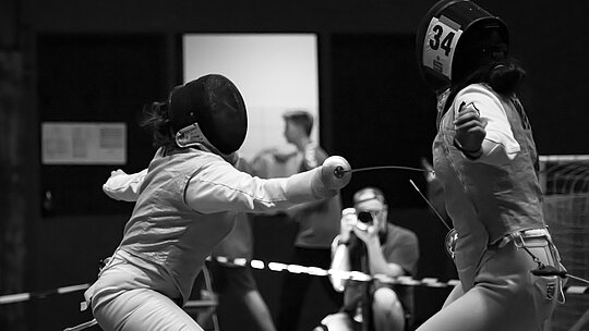 A foto em preto e branco mostra duas esportistas durante o combate de esgrima. Uma está atacando a outra, conseguimos ver bem sua máscara e arma posicionada no corpo da adversária - que está de costas.