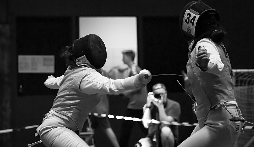 A foto em preto e branco mostra duas esportistas durante o combate de esgrima. Uma está atacando a outra, conseguimos ver bem sua máscara e arma posicionada no corpo da adversária - que está de costas.