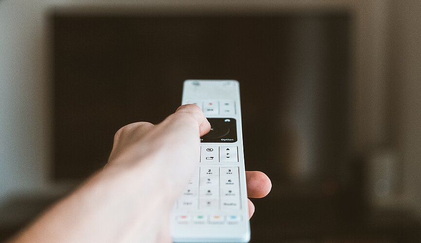 Na imagem a mão de uma pessoa segura um controle branco, mirando diretamente para TV. Somente o controle está focado. 