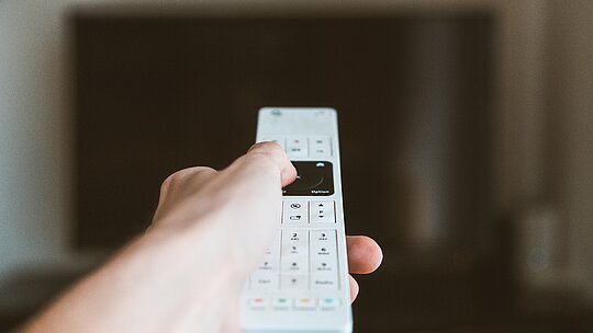 Na imagem a mão de uma pessoa segura um controle branco, mirando diretamente para TV. Somente o controle está focado. 