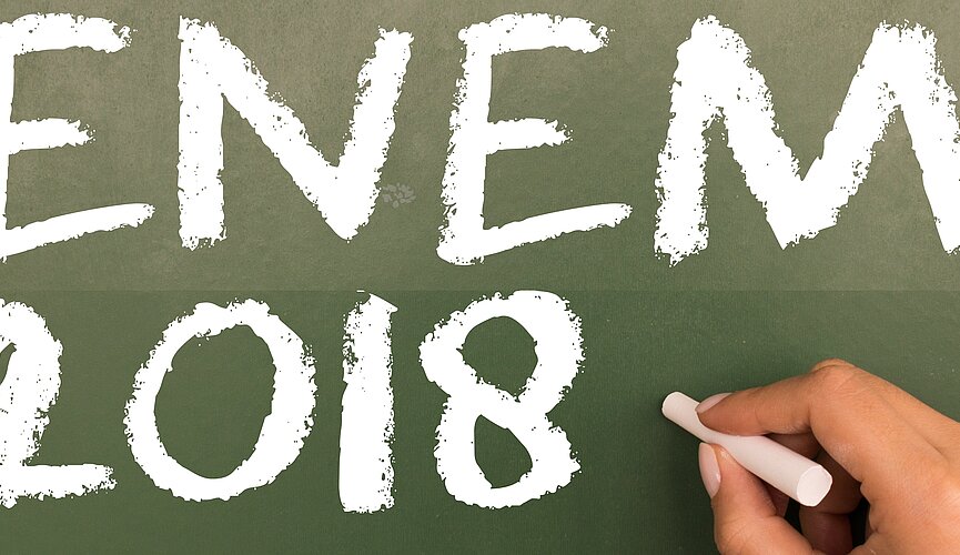 Mão segurando giz com a inscrição "Enem 2018" no quadro negro