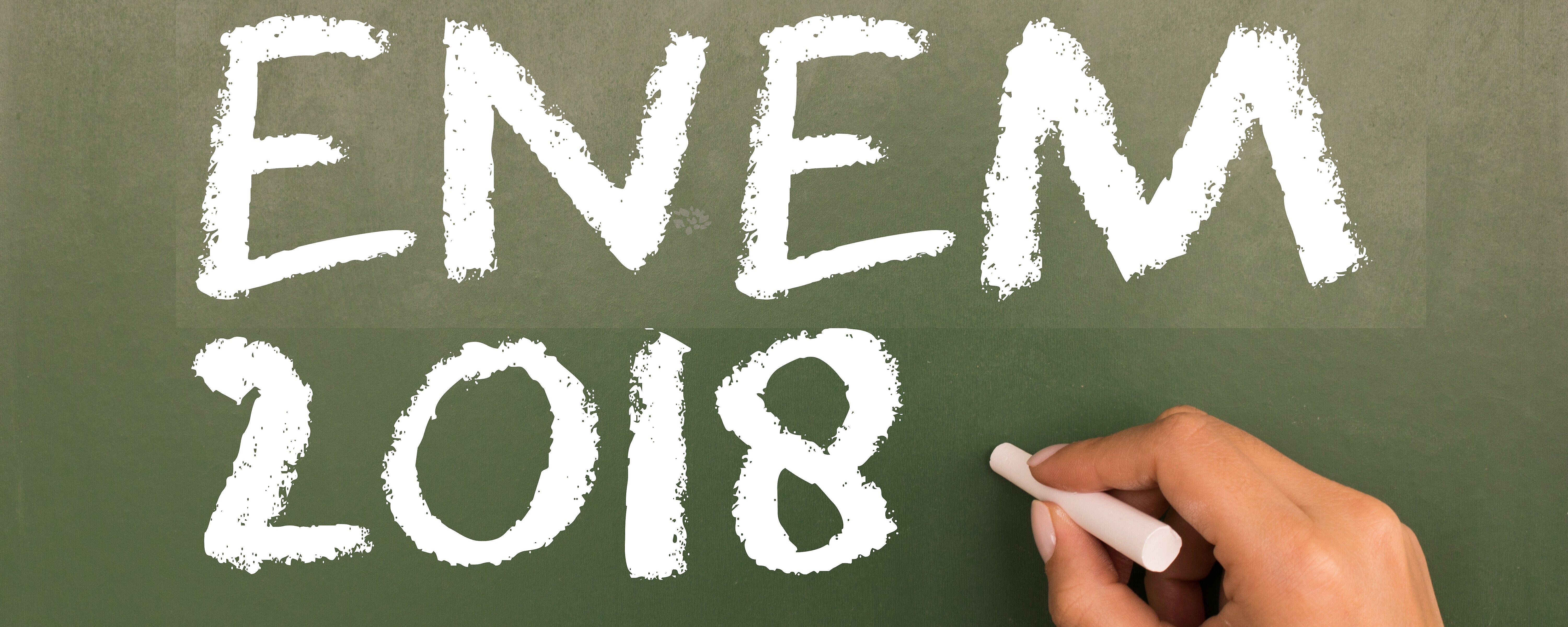 Mão segurando giz com a inscrição "Enem 2018" no quadro negro