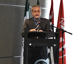 Pró-reitor está falando no pulpito em cima do palco do auditório do MackGraphe. Atrás estão algumas bandeiras.