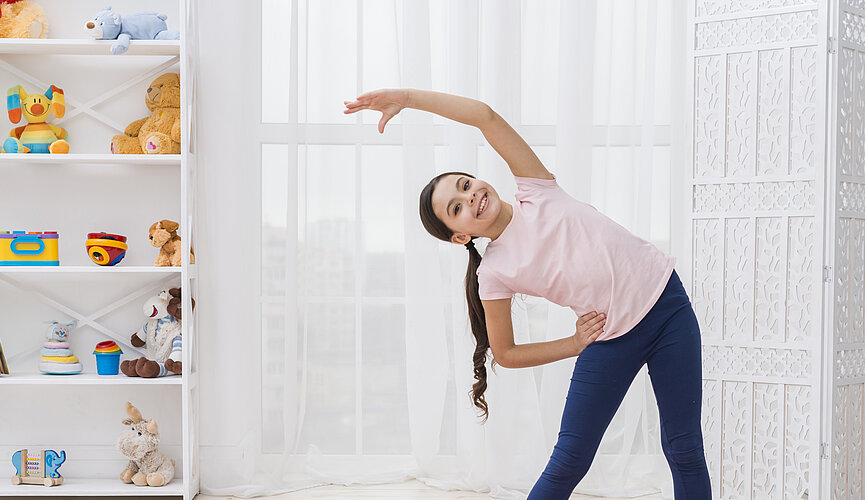 Uma menina de cabelos morenos está fazendo um alongamento, dentro do seu quarto. Logo atrás dela, tem uma janela com cortinas e também uma estante, cheia de brinquedos e bichos de pelúcia. A foto é bem clara e feliz.