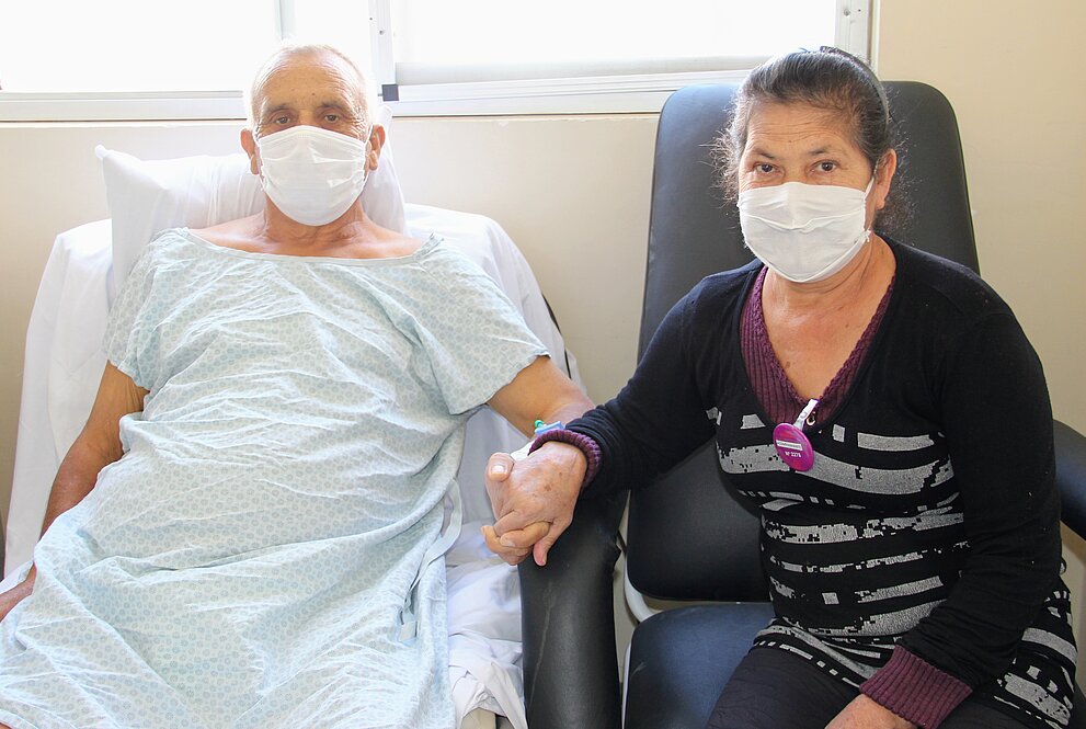 O paciente Leonardo, com máscara e camisola cirúrgica, está de mãos dadas com sua esposa, Sirley Machado Ogrodovski, também de máscara. Ambos estão sentados.  