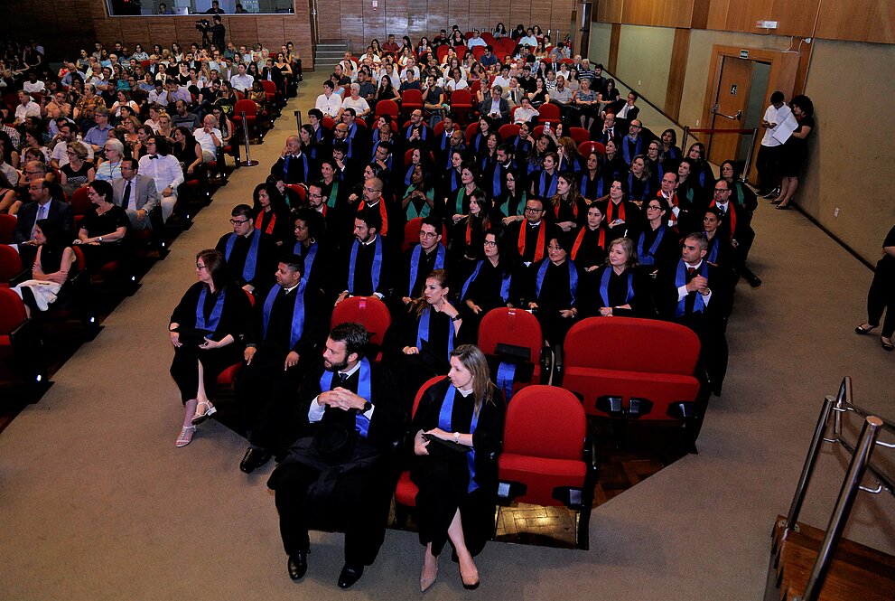 Alunos doutorandos sentados na plateia do auditório com togas de formatura.