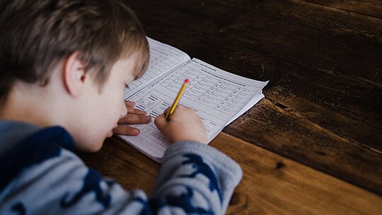A foto mostra, de cima para baixo, um menino estudando. Ele está com um lápis na mão e um livro sobre a mesa. 