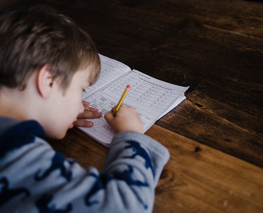 A foto mostra, de cima para baixo, um menino estudando. Ele está com um lápis na mão e um livro sobre a mesa. 