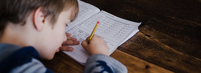 A foto mostra, de cima para baixo, um menino estudando. Ele está com um lápis na mão e um livro sobre a mesa. 
