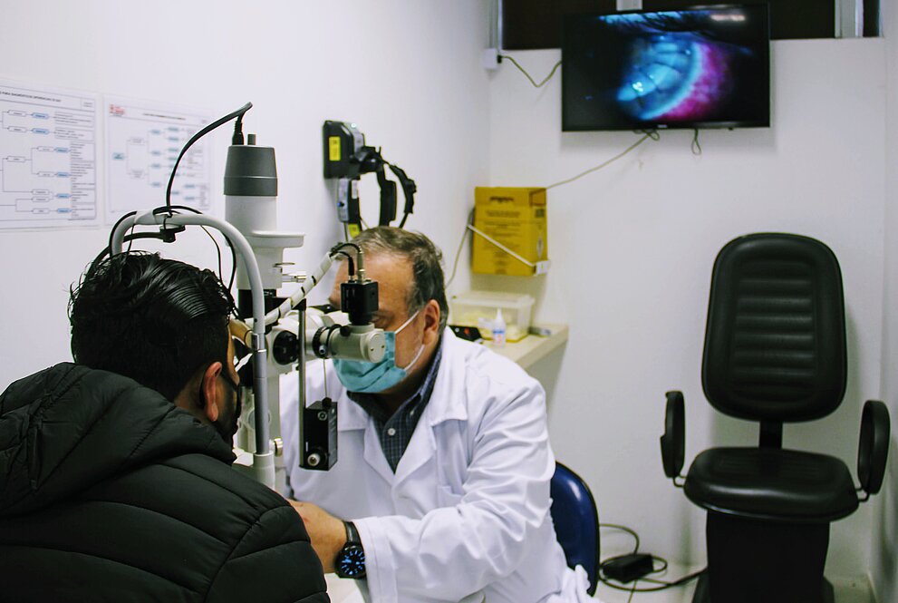 Num consultório de oftalmologia, médico de jaleca examina os olhos de um paciente que veste jaqueta preta. O médico está de frente na imagem e o paciente de costas para o fotógrafo