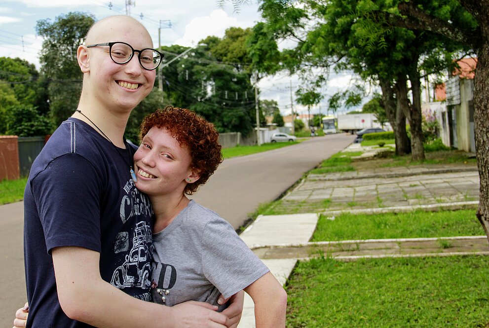 um homem branco, sem cabelos e de óculos abraçado com uma garota branca de cabelos vermelhos encaracolados, ao fundo, uma rua tranquila de subúrbio com árvores e gramados