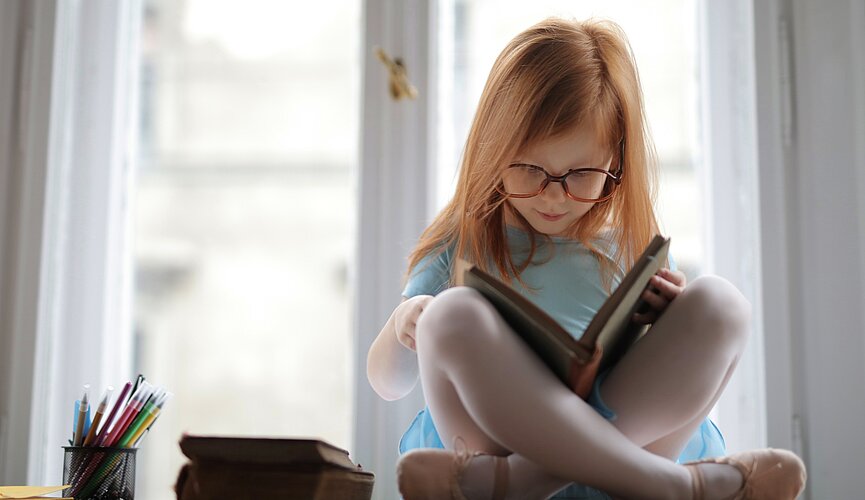 Na foto, uma menina ruiva, de cabelos lisos, está sentada em cima de uma mesa, lendo um livro, com outros livros do lado e muitos lápis em um potinho. A foto foi tirada em frentre a uma janela.