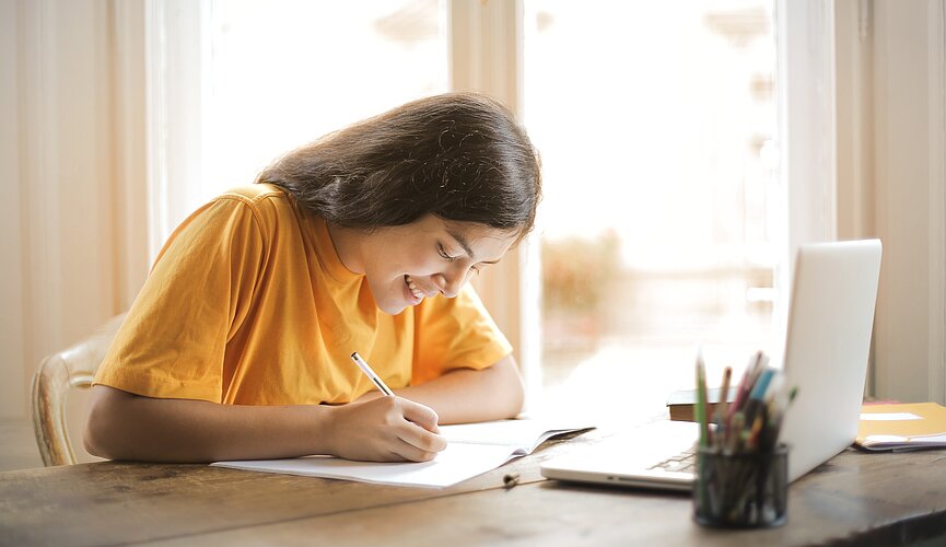 Imagem mostra uma menina escrevendo em um folha e um notebook a frente