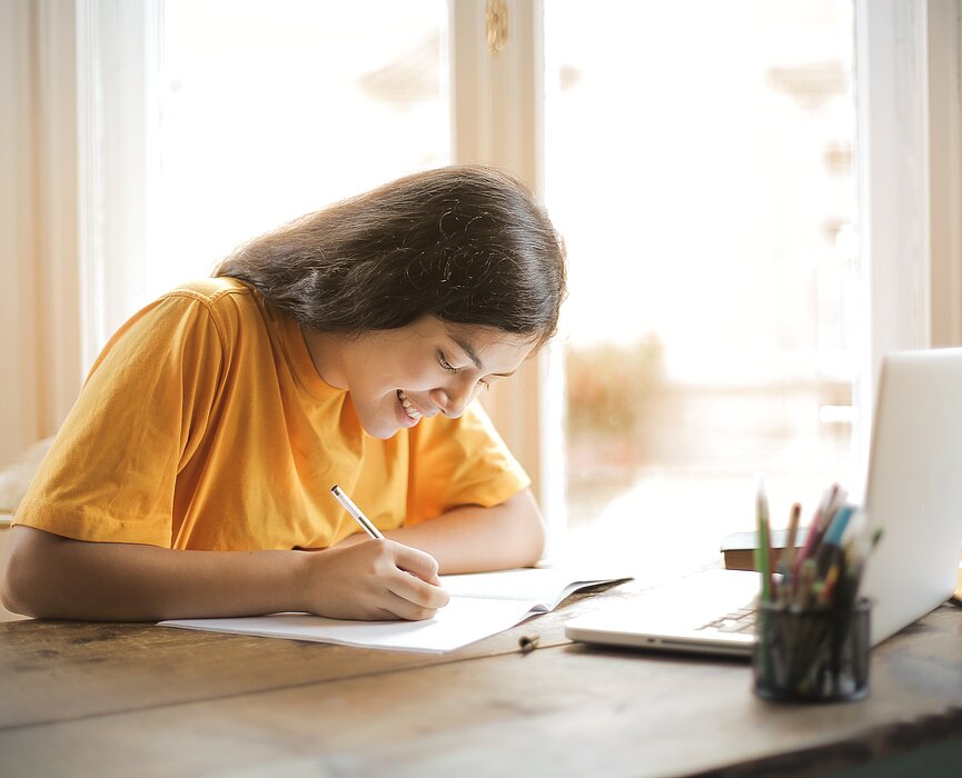 Imagem mostra uma menina escrevendo em um folha e um notebook a frente