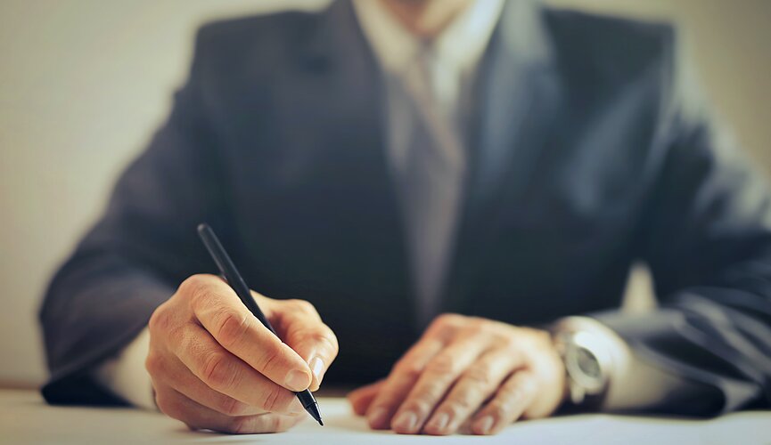 Imagem com um homem de terno escrevendo com caneta na folha