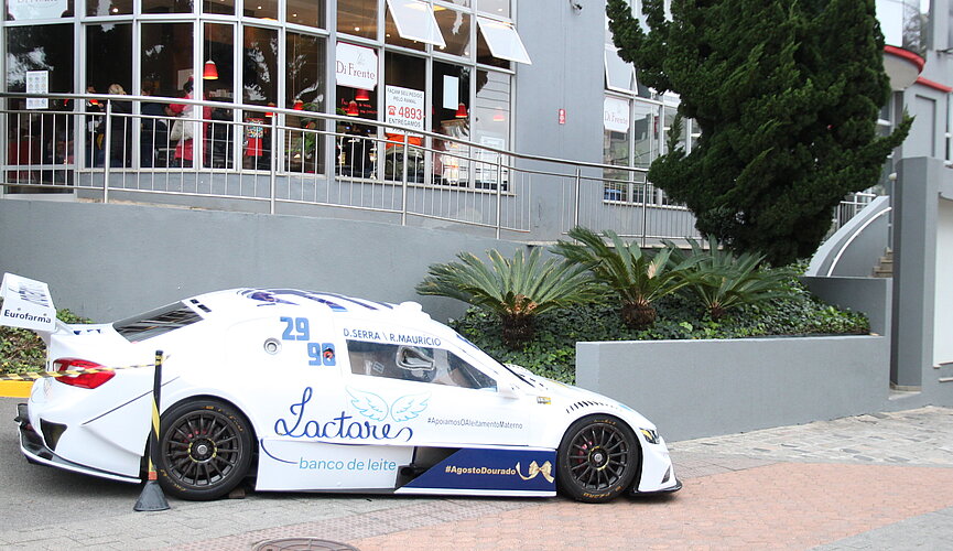 Carro branco da Stock Car estacionado em frente ao HUEM. Atrás pode-se ver o prédio cinza com logotipos em vermelho