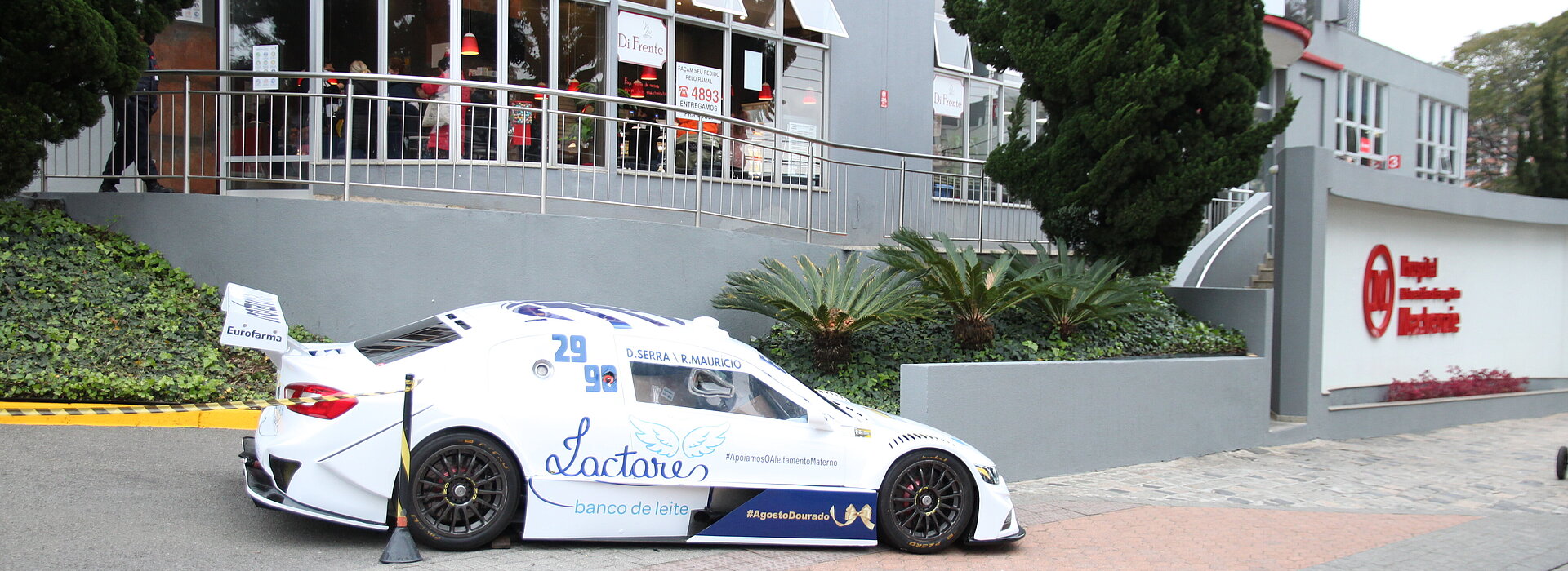 Carro branco da Stock Car estacionado em frente ao HUEM. Atrás pode-se ver o prédio cinza com logotipos em vermelho