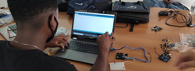 Na foto, um homem de costas usa um laptop, que está em cima de uma mesa, cheia de ferramentas, cabos e itens utilizados no curso de eletricidade.