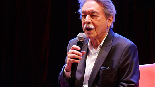 A foto mostra o arquiteto Paulo Mendes da Rocha em um evento do Mackenzie, no ano de 2017. Ele está com um blazer e segurando um microfone, sentado em uma poltrona vermelha.